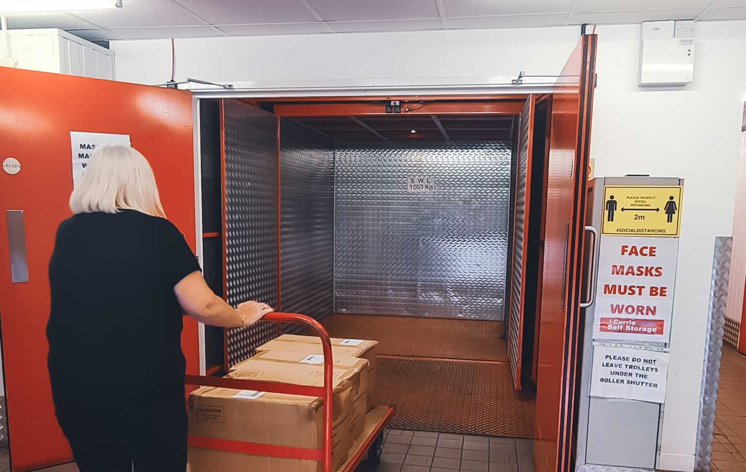 A woman is pushing a pallet trolley loaded with cardboard boxes into a lift.