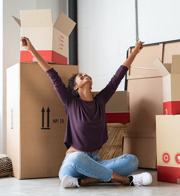 A smiling woman surrounded by cardboard boxes is sat on the ground with her hands in the air.