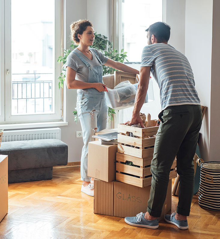 A couple are stood talking to each other as they move boxes around their home.