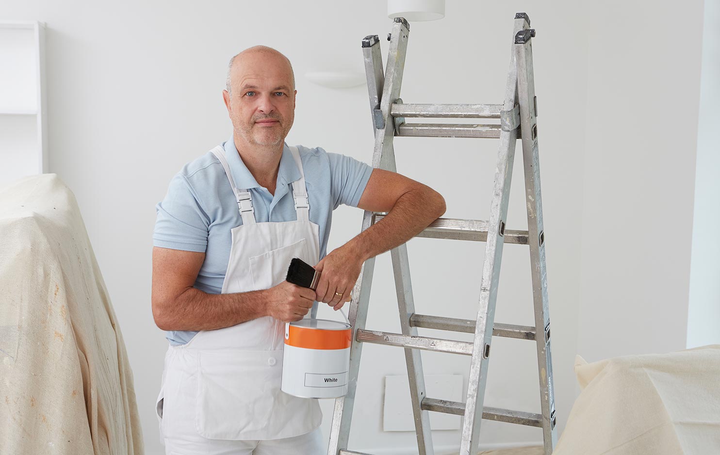 A tradesman is holding a pot of paint and paintbrush whilst leaning against a step ladder.