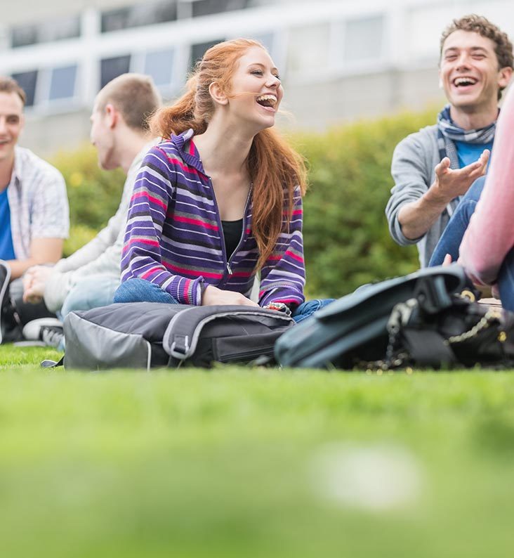A group of friends are sat on the grass and laughing.