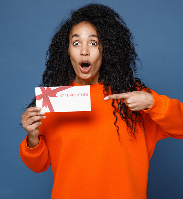 A woman is holding a gift voucher and is pointing to it with her other hand.